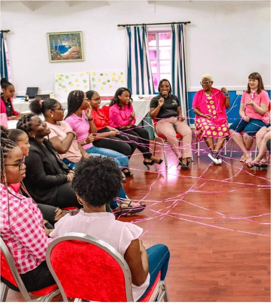 group teenage girls sitting in a circle engaged in a workshop, holding on to a piece of string that is woven with the pieces of string held by the other girls and the two women leading the workshop.